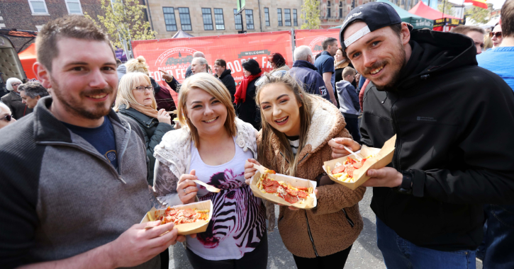 Two men and two women smiling at camera holding pizza slices and chips at Bishop Auckland Food Festival.